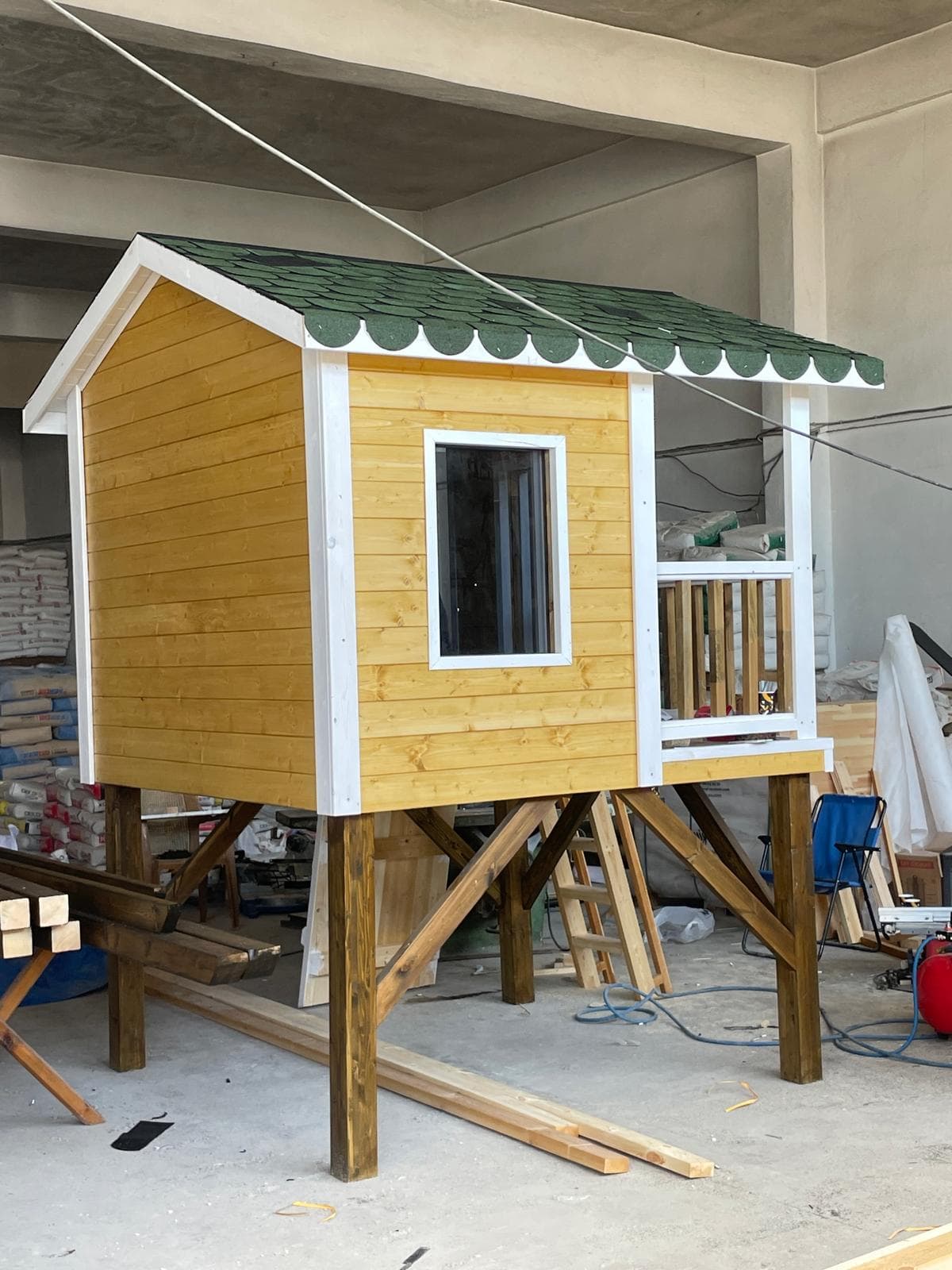 Sarı koruyucu boyalı, verandalı ve yükseltilmiş ayaklı ahşap çocuk oyun evi